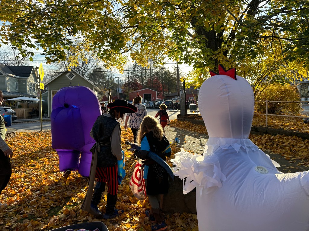 Children dressed in costumes participate in a festive Halloween event, surrounded by colorful autumn leaves and inflatable decorations.